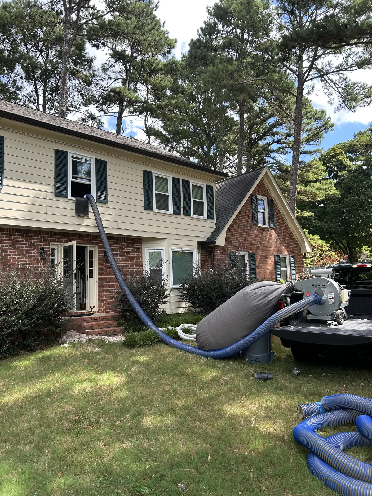 Two-story home with an insulation removal hose running from an upstairs window to vacuum equipment on a truck